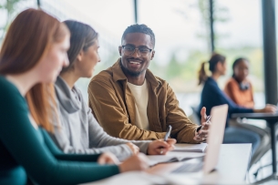 A small group of university students are seen sitting side by side at a desk in class as they work on an assignment. They each have books open in front of them as they collaborate and share ideas.