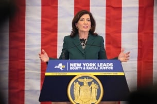 Light-skinned woman with dark hair at a podium in front of U.S. flag