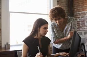 A woman in an office setting stands over a younger woman looking at a computer, helping her.