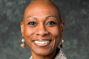 Antoinette "Bonnie" Candia-Bailey, a Black woman with a bald head, wearing a textured jacket and a statement necklace with matching earrings, smiles in a professional headshot.