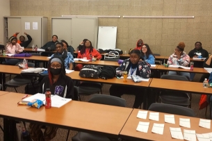 A group of young people sit at tables in a classroom doing work and smiling at the camera.