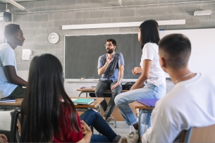 A male teacher leads four students, sitting in a circle and two of them at desks while two of them are sitting on top of desks.