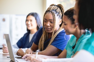 A diverse group of women in scrubs sit at a desk and look at a laptop.