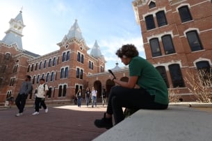 A student with headphones sits near a brick building on a college campus.
