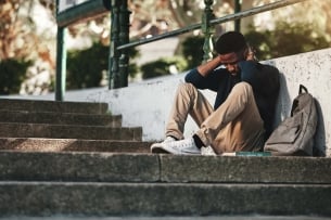 A student sits on stairs next to a backpack, looking frustrated and sad.