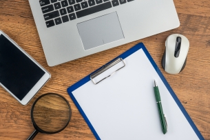 Desktop with computer, iphone, magnifying glass, and clipboard with pen and blank paper