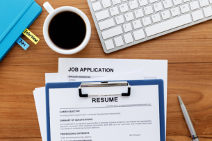 A clipboard holding an applicant's résumé sits on a desk above a job application. From left to right, a blue folder bearing tabs that say "interview tips," a cup of coffee and a keyboard are also visible on the desk.