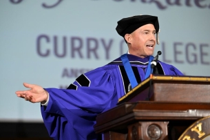 Curry College President Jay Gonzalez wears academic robes and gestures with his arms at a podium with “Curry College” projected in purple letters behind him. 