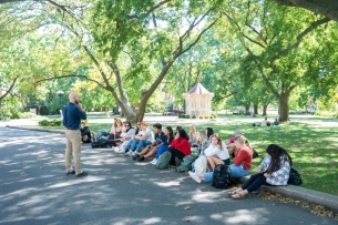 A Belmont University faculty member teaches a class outside to a row of students sitting on a curb with a green lawn and trees behind them.
