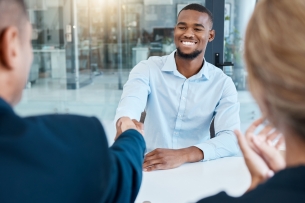 A young man shakes hands with a person across the table, smiling, in an office.