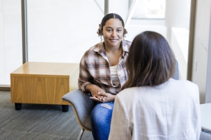 A female college student in a plaid shirt sits across the desk from a professional providing support.