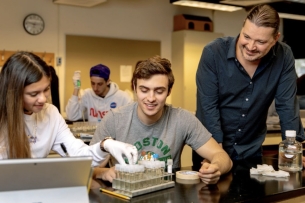 Mark Sarvary stands next to two seated students working with test tubes in a lab course setting, smiling.