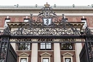 The front gate of Barnard College. A brick building is in the background with a wrought iron fence in the front, which says "Barnard College of Columbia University"