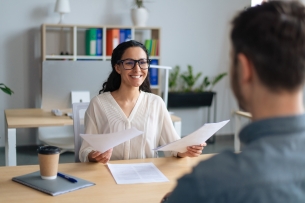 Woman at office desk in front of bookcase smiles as she talks to young man, whose head you see from the back