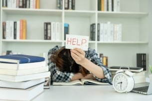 A student sits slumped over an open book with their head on their arm atop the open book, surrounded by other textbooks and a clock, and holding up a sign that says "help" in all capital letters.