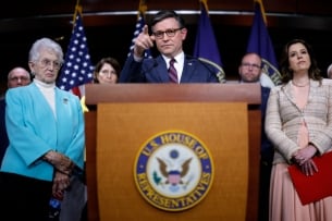 Speaker Mike Johnson points a finger while standing behind a podium. Virginia Foxx and Elise Stefanik stand next to him.