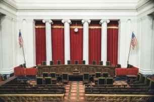 A photo of the interior of the U.S. Supreme Court chambers, showing a curved table with nine chairs in front of a set of five long red curtains hanging between four white columns.