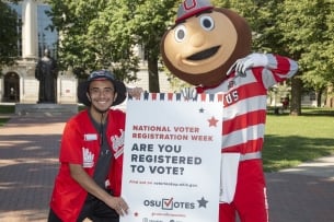 A student standing beside Ohio State University mascot Brutus Buckeye holding a sign asking students if they are registered to vote.
