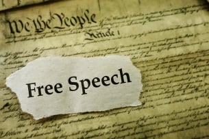 A cutout of a piece of paper with the words "Free Speech" laid atop a sepia-toned copy of the U.S. Constitution.