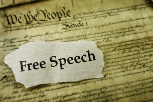 A cutout of a piece of paper with the words "Free Speech" laid atop a sepia-toned copy of the U.S. Constitution.