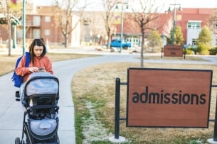A woman pushes a baby stroller on a college campus next to a sign that says "admissions." She is texting on her phone as she walks