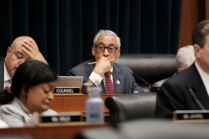 A light-skinned Black man with gray hair wearing a suit and tie sits at a desk