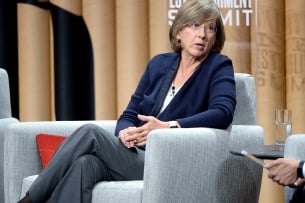 A woman with glasses wearing in a blue sweater sits onstage, leaning on the arm of her chair as she speaks to someone next to her.