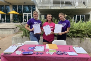 Three young woman stand behind a table holding voter registration forms.