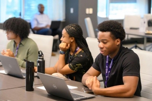 Three students sit at a conference table working on laptops.