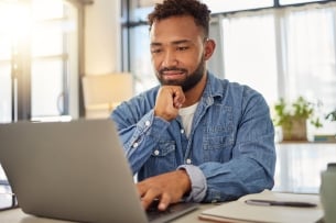 An adult man works at a laptop with a notebook and pen beside him.