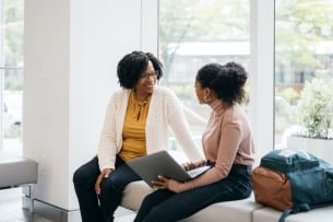 A stock photo of a Black woman professor speaking to an engaged student holding a laptop. The professor is smiling and the two appear to be in a good conversation in front of a wall of windows.