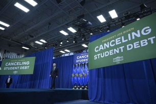 President Biden walks on a stage flanked by two signs that say "Canceling Student Debt" in white text.