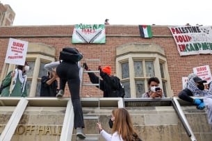 Pro-Palestinian protestors trying to set up a solidarity encampment at the University of California, Los Angeles's Kerckhoff Hall May 23. One protester climbs a ladder to what appears to be a roof to join other protesters; signs in the background espouse support for Palestine.