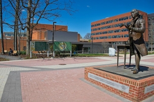 A statue of George Mason sits at the forefront of this image of George Mason University's campus. University buildings can be seen in the background.