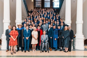 A group of University of Austin students in blue robes stand on stairs behind faculty and Texas governor Greg Abbott.