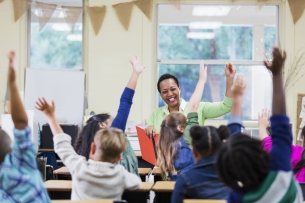 An African American teacher, a mature woman in her 40s, sitting in front of her class of elementary school students, 6 and 7 years old, reading a book. She has asked a question and most of the children are raising their hands. They are in first grade or second grade.