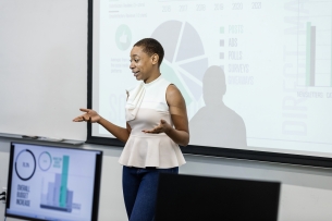 An adult female engineer gestures while presenting data to her peers in a conference room.