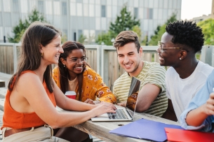 Multiethnic group of happy and smiling young students preparing for an exam and doing homework, sharing different ideas using their laptops in positive and friendly communication on campus.