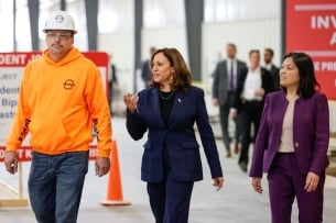 A man in a construction hat and neon vest walks next to Vice President Kamala Harris and acting labor secretary Julie Su. 