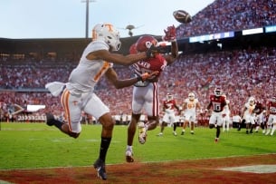 A football player in white and orange tries to catch a ball but is blocked by a player in red. They are on green turf in a crowded stadium.