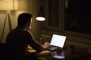 A male student, photographed from behind, works alone on a laptop at night; the room is dark, save for the illumination provided by two lights and the glow of the laptop screen.