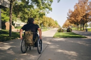 An adult disabled man, attending college, with a camo backpack hanging from his wheelchair.