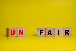 Six wooden alphabet blocks are juxtaposed against a bright yellow background in this illustration of the concept of fairness and unfairness. Two of the blocks read "UN" and are separated with a space from four more blocks that say "FAIR."