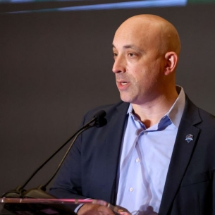 Jonathan Greenblatt, a light-skinned man with a bald head, wearing a blue collared shirt and a blue blazer, stands a podium