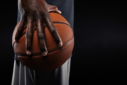A close-up photo of a Black athlete’s hand holding a basketball, against a black background..