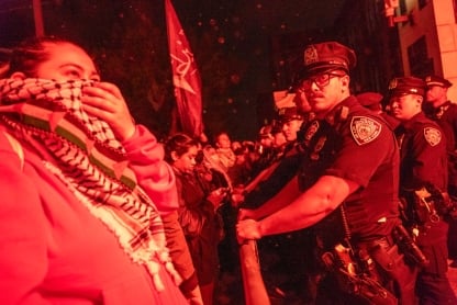 In this red-toned image, pro-Palestinian protesters stand face-to-face in a confrontation with police at the City College of New York on April 30. In the foreground, a protester holds a keffiyeh to her face as a mask while a male police officer looks on.