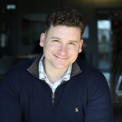 Michael Mindzak, a light-skinned man with curly dark hair, wearing a blue zip-up sweater over a checked collared shirt