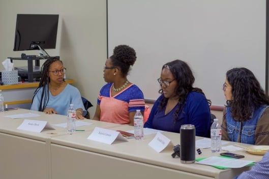 Four students participating in a panel are seated at a table, where they are speaking with and engaging with one another.