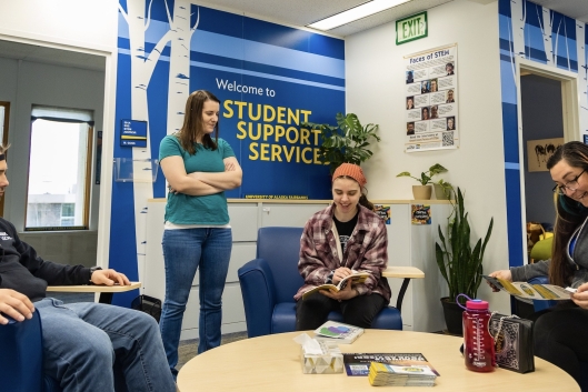 Three seated people in casual clothes speak with a woman standing in a warm-looking campus office space. A large wall sign says "Welcome to Student Support Services."