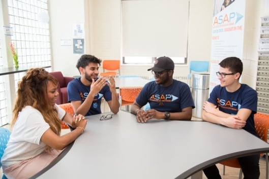 Four students sit around a table talking. Three of them are wearing navy blue shirts that read "ASAP" in blue and orange letters. 